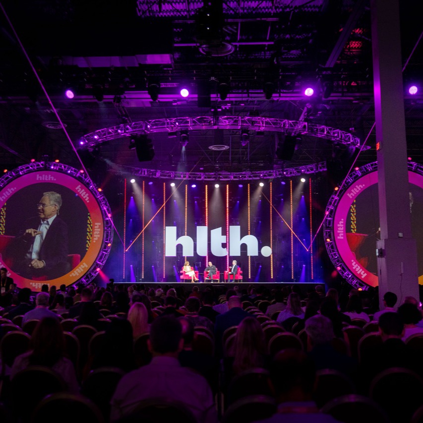 The HLTH main stage, with a panel seated in front of the hlth. screen and a full audience
